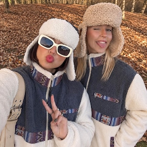 May include: Two zip-up fleece jackets featuring a dark charcoal grey chest panel, cream-colored sleeves and lower body, and a colorful woven geometric pattern trim. Each jacket includes a matching sherpa-lined trapper hat with ear flaps.