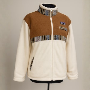 Puede incluir: Chaqueta de forro polar crema y marrón con cremallera completa y cuello alto. La chaqueta tiene la parte superior del pecho y los hombros marrones, con mangas y cuerpo color crema. Incluye detalles estampados y un pequeño parche.