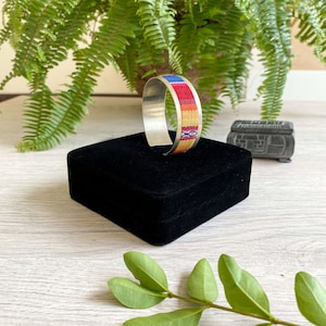 May include: A silver bangle bracelet with a colorful woven band. The band features red, yellow, blue, and white stripes. The bracelet is displayed on a black velvet box.