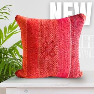 May include: A square, textured pillow in shades of red and orange. The pillow features a central floral design and is placed on a white surface. The word "NEW" is visible in the background.