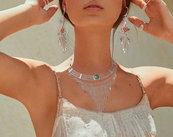 May include: A woman wearing a white, crocheted dress with a silver necklace and earrings. The necklace has a green stone pendant and the earrings have pink stones.