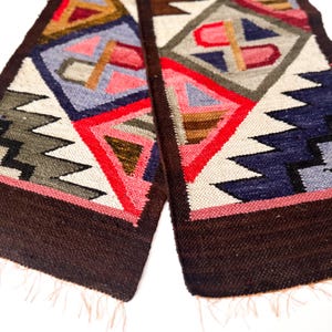 Op de afbeelding: Close-up van twee rechthoekige geweven textielstukken met geometrische patronen. De ontwerpen bevatten driehoeken, ruiten en zigzags in rood, blauw, roze en bruin. De randen zijn donkerbruin met franjes.