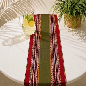 May include: A colorful table runner with a central olive green stripe and red borders. The runner is decorated with vertical stripes in various colors. A glass pitcher with lemon slices and a fern in a woven basket are also visible.