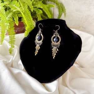 May include: A pair of gold dangle earrings with a dark blue gemstone in the center of each earring. The earrings have a delicate, intricate design.