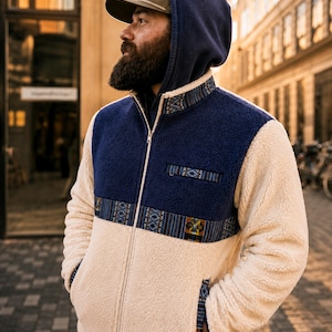 May include: A zip-up fleece jacket featuring a cream-coloured body and sleeves with a dark blue upper chest panel. The jacket includes a patterned trim along the chest, collar, and pocket edges, a full-length front zip, and a stand-up collar.