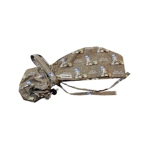 May include: A brown surgical scrub hat with a repeating pattern of R2-D2 robots and the text "Small but Mighty". The hat has a tie on the back and a small pouch attached to the side.