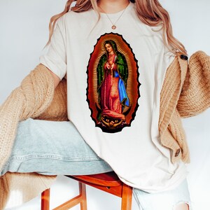 May include: A white t-shirt with a colorful image of the Virgin Mary on the front. The image is surrounded by a gold halo and has a blue and green robe.