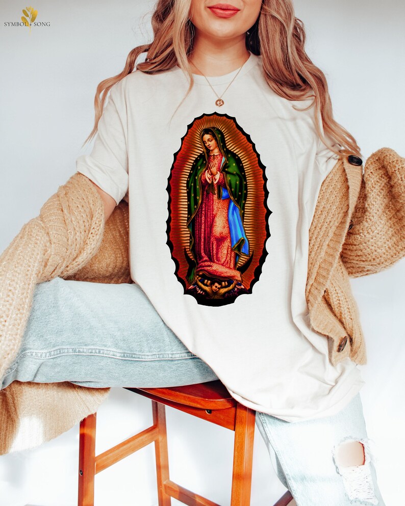 May include: A white t-shirt with a colorful image of the Virgin Mary on the front. The image is surrounded by a gold halo and has a blue and green robe.