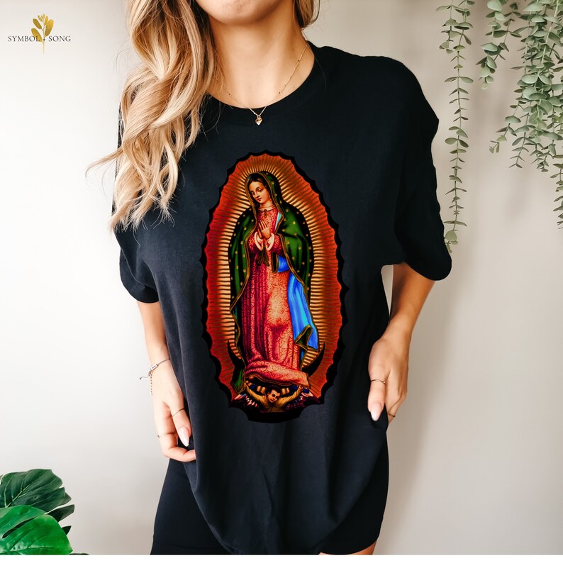 May include: Black t-shirt with a colorful image of Our Lady of Guadalupe, a religious figure in a blue and green robe, standing in a golden halo.
