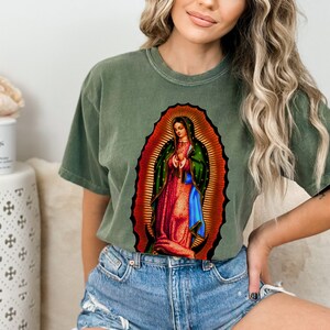 May include: A green t-shirt with a graphic print of Our Lady of Guadalupe, a religious image of the Virgin Mary.