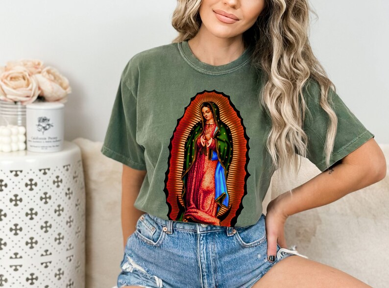 May include: A green t-shirt with a graphic print of Our Lady of Guadalupe, a religious image of the Virgin Mary.