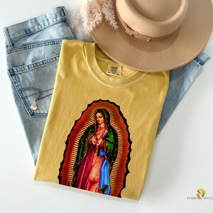 May include: A mustard yellow t-shirt with a graphic print of Our Lady of Guadalupe. The t-shirt has a crew neck and short sleeves. The graphic is a colorful depiction of the Virgin Mary in a blue and green robe, with a gold halo and a crown of stars.