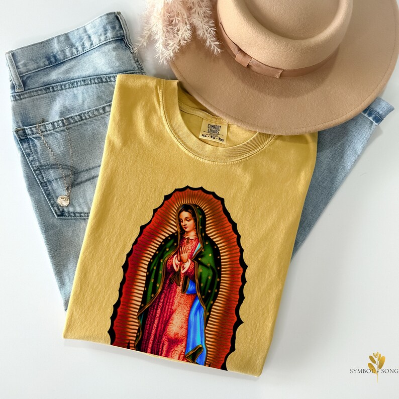 May include: A mustard yellow t-shirt with a graphic print of Our Lady of Guadalupe. The t-shirt has a crew neck and short sleeves. The graphic is a colorful depiction of the Virgin Mary in a blue and green robe, with a gold halo and a crown of stars.