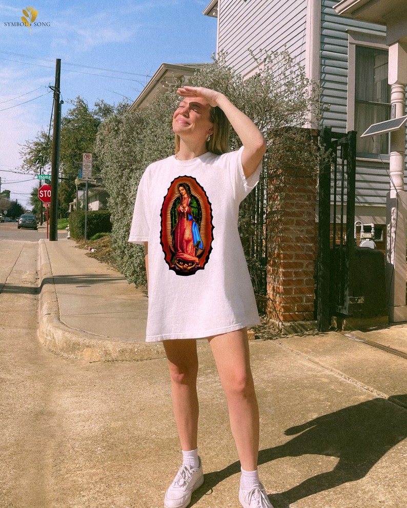 May include: A white oversized t-shirt with a colorful image of the Virgin Mary on the front. The person wearing the shirt is standing on a sidewalk in front of a house.