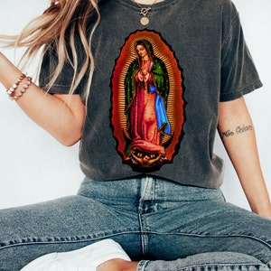May include: A gray t-shirt with a colorful image of the Virgin Mary. The image is surrounded by a gold halo and the Virgin Mary is wearing a blue robe and a red dress.