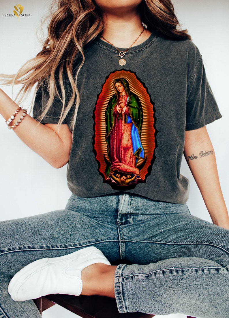 May include: A gray t-shirt with a colorful image of the Virgin Mary. The image is surrounded by a gold halo and the Virgin Mary is wearing a blue robe and a red dress.