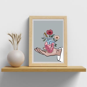 Puede incluir: Una impresión artística enmarcada que presenta una ilustración estilizada de un corazón humano con flores. El corazón es sostenido en una mano, con un fondo gris claro. El marco es de madera. Un jarrón con trigo está en una estantería de madera.