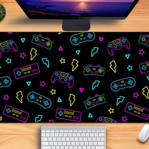 May include: A black gaming mouse pad with a neon design featuring video game controllers, lightning bolts, and other symbols. The design is in shades of pink, blue, yellow, and green.