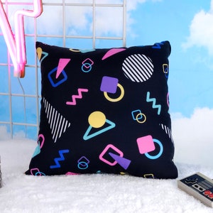 May include: A black cushion with a geometric pattern in pink, blue, yellow, and purple. The pattern includes circles, squares, and zigzags.