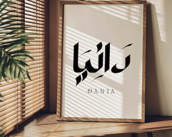 May include: A framed print with the Arabic word "Dania" written in black calligraphy. The word is centered on a white background with a light brown wooden frame.