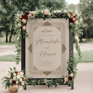 May include: A welcome sign for a wedding with a gold ornate frame and the text "Welcome to the Nikkah of Moustafa and Noura 30.12.2022". The sign is decorated with flowers.