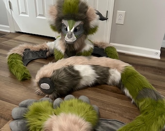 May include: A multi-colored furry costume, featuring a headpiece, arms, tail, and hand covers. The costume has a green, gray, white, and pink color scheme. The headpiece has a detailed face with a gray snout and white teeth. The costume is designed to resemble a fantasy creature.
