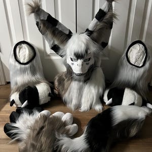 Op de afbeelding: Een harig kostuum met een wit, zwart en bruin kleurenschema. De set bevat een hoofddeksel met grote, patroonoren, twee pootachtige handhoezen, twee voetbedekkingen en een lange staart. Het hoofddeksel heeft een wit schedelachtig masker.
