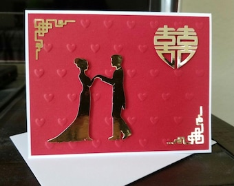 May include: A red wedding card with embossed hearts and gold foil accents. The card features a silhouette of a couple holding hands, a double happiness symbol, and decorative corner designs. The card is on a white envelope.