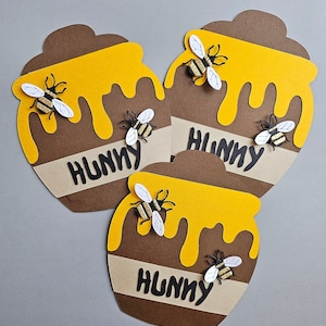 May include: Three paper cutouts of honey pots with yellow honey dripping down. Each pot is brown with a tan band that reads "HUNNY" in black letters. Three-dimensional bees with white wings and gold and black bodies are attached to the pots.