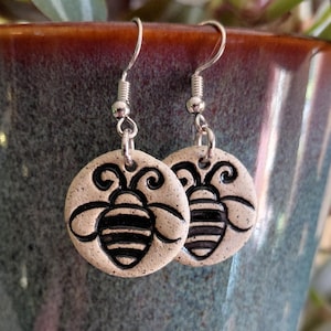 May include: A pair of handmade earrings featuring a circular, beige pendant with a black bee design. The earrings have silver-colored hooks and are displayed against a blue-green mug with a brown rim, with green and purple foliage in the background.