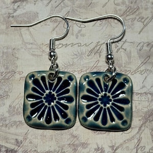 May include: A pair of square ceramic earrings with a floral design. The earrings feature a dark blue flower pattern on a light blue background, with silver-colored ear wires. The earrings are a unique handmade accessory.