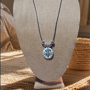 May include: A black cord necklace with silver beads and a white ceramic pendant featuring a hand-drawn bee design.