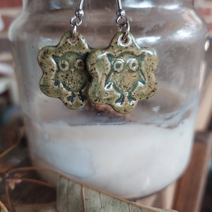 May include: A pair of green and brown ceramic owl earrings with silver hooks. The owls have a speckled glaze and are in a starburst shape.