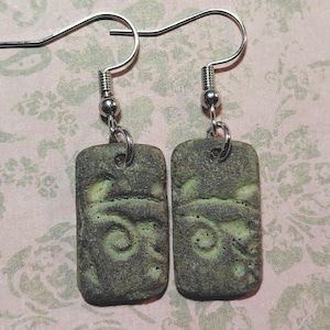May include: A pair of handmade earrings with a rectangular, textured design. The earrings feature a dark green and brown color scheme with a swirling pattern. The ear hooks are silver-colored.