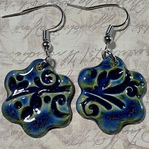 May include: A pair of ceramic earrings with a floral design. The earrings are a mix of blue and green with a glossy finish. The hooks are silver-colored.
