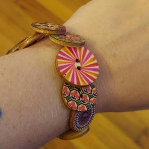 May include: A wooden button bracelet featuring a variety of colorful, patterned buttons. The bracelet includes buttons with floral, striped, and geometric designs in shades of pink, orange, and purple. The bracelet is worn on a wrist.