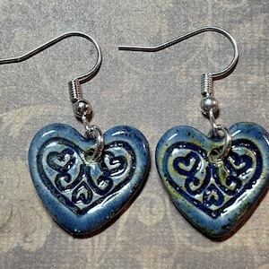 May include: A pair of heart-shaped earrings with a blue glaze and intricate dark blue patterns. The earrings have silver-colored hooks and are displayed against a textured background. The heart pendants are approximately 1 inch wide.