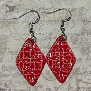 May include: A pair of red diamond-shaped earrings with a floral pattern. The earrings have a glossy finish and are suspended from silver-colored ear wires. The design features a raised floral pattern on a red background.