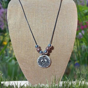 May include: A black cord necklace with a silver pendant featuring a detailed design of two hearts surrounded by foliage. The necklace has brown and silver beads.