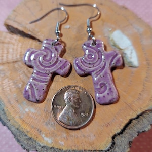 May include: A pair of purple cross earrings with a swirl design. The earrings are made of ceramic and have silver hooks.