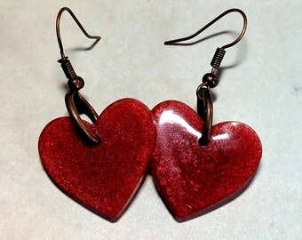 May include: A pair of heart-shaped earrings. Each earring features a vibrant red heart with a glittery texture, suspended from a bronze-colored hook. The earrings are designed for pierced ears and are suitable for everyday wear or special occasions.