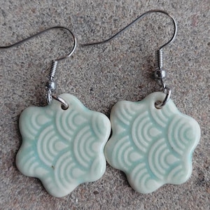 May include: A pair of ceramic earrings with a flower shape and a light blue and white wave pattern. The earrings have silver-colored ear wires.