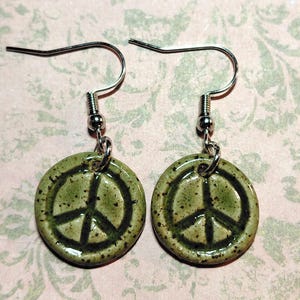 May include: A pair of ceramic earrings featuring a peace symbol. The earrings have a circular, olive-green base with a black peace sign. The ear wires are silver-colored.