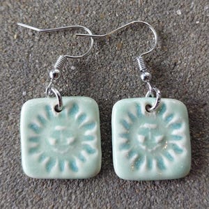 May include: A pair of square, light green ceramic earrings with a sun design in a darker shade of green. The earrings have silver-colored hooks. The sun design is embossed on the surface of each earring.