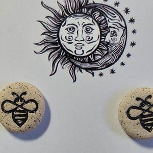 May include: Two round, beige stone earrings with a black bee design. The earrings are displayed on a white background with a black and white illustration of the sun, moon, and stars.