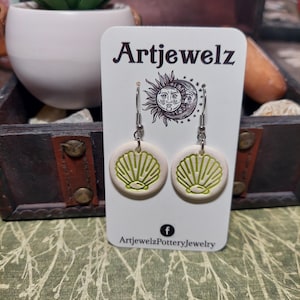 May include: A pair of white ceramic earrings with a green seashell design. The earrings are on a white card with the text "Artjewelz" and "ArtjewelzPotteryJewelry" on it.