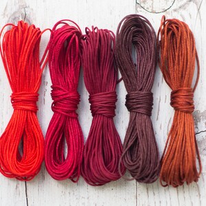 Könnte beinhalten: Fünf Bündel farbiger Kordeln in Rot-, Burgunder- und Brauntönen. Die Kordeln sind fest gewickelt und scheinen aus einem glatten, glänzenden Material zu bestehen. Die Kordeln sind auf einer weißen, verwitterten Holzoberfläche angeordnet.