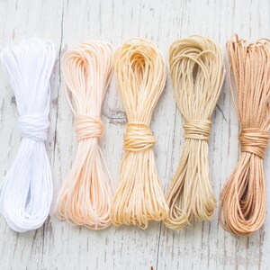 Könnte beinhalten: Fünf Spulen mit Kordel in verschiedenen Beige-, Weiß- und Brauntönen. Die Kordeln sind horizontal auf einer weißen Holzoberfläche angeordnet.