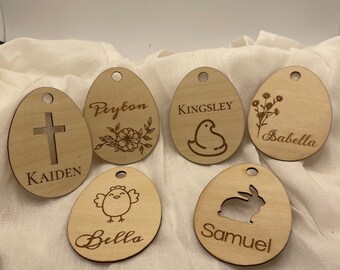 May include: Six wooden Easter egg ornaments with different designs and names. The names are Kaiden, Peyton, Kingsley, Isabella, Bella, and Samuel. Each ornament has a different design, including a cross, flowers, a chick, a bunny, and a bird.