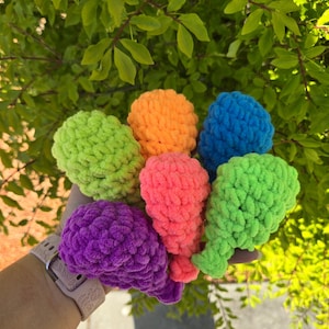 May include: A collection of colorful, handmade crochet water balloons in shades of green, orange, blue, pink, purple, and green. The soft, textured yarn creates a playful and tactile appearance. The balloons are held against a backdrop of green foliage.